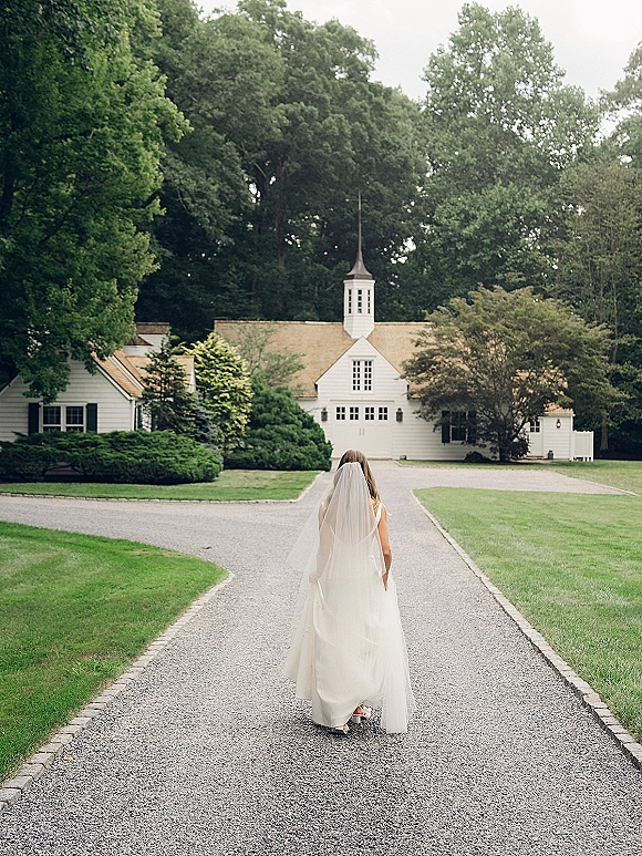 Bride walking away down a gravel driveway in a wedding dress with a long veil, heading toward a white chapel amid trees and lawn