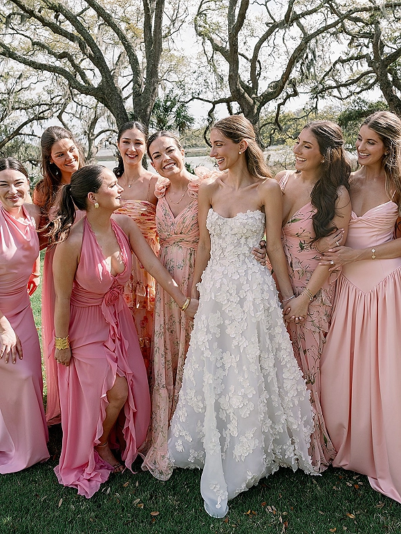 Bridesmaids group photo of bride with bridesmaids in pink and floral dresses, laughing on a lawn by oak trees with hanging moss and river