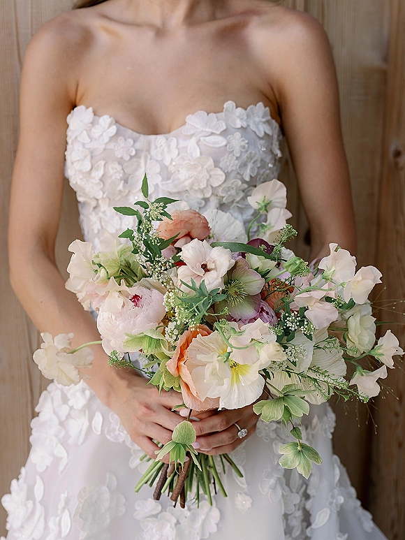 Bridal bouquet in pastel wedding bouquet tones of blush and white with sweet peas and greenery, held against a strapless lace dress by a wood wall