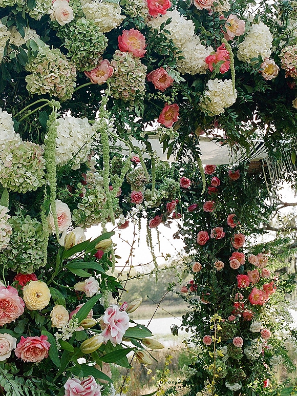 Floral ceremony arch with garden rose clusters and flowing fabric draping, framed by trees and a lakeside outdoor landscape backdrop
