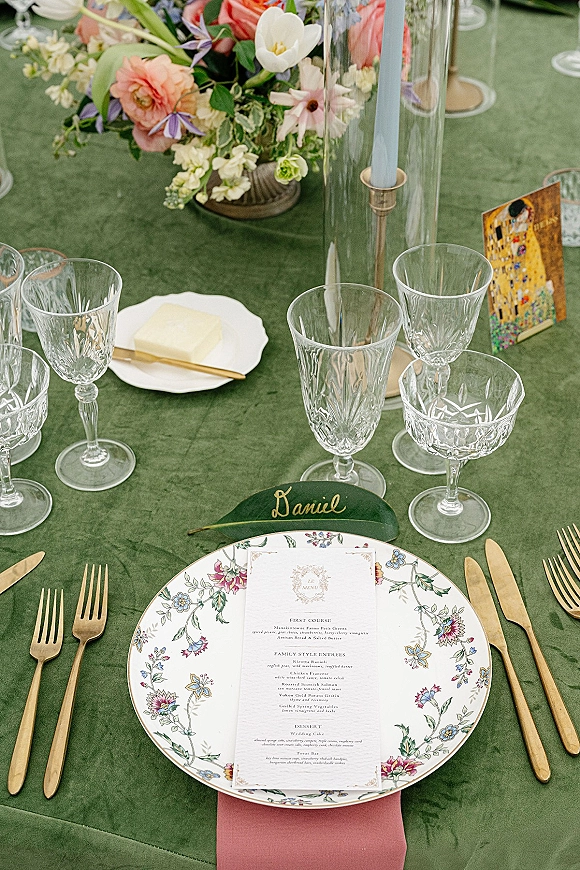 Reception tablescape with green wedding tablecloth, floral centerpiece, blue taper candle in glass hurricane, gold flatware and crystal stemware