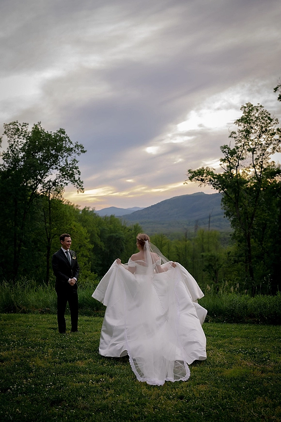 First look moment as bride in wedding dress and veil walks toward groom in black suit across a grassy field with mountain sunset backdrop