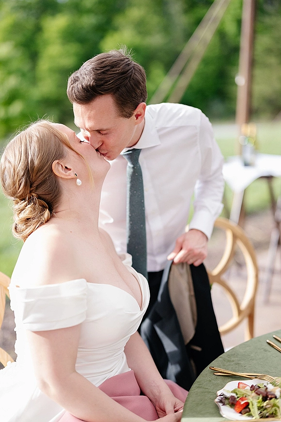 Wedding kiss as the bride and groom kiss at an outdoor tent reception table, bride in off the shoulder dress beside green linens and salad plate