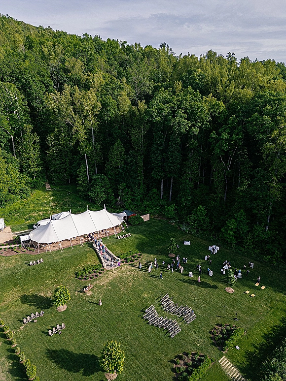 Outdoor wedding venue with a sailcloth tent, ceremony chairs and cocktail tables set on a grassy field bordered by forest trees