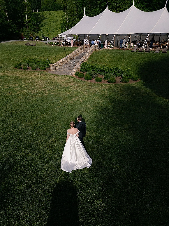 Couple portrait from above of bride and groom walking across a lawn, her wedding dress train trailing toward a white tent with lights