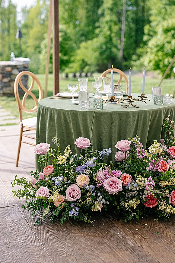 Reception tablescape with a green wedding tablecloth, brass taper candles, crystal glassware, and roses on an outdoor patio by trees and a stone wall