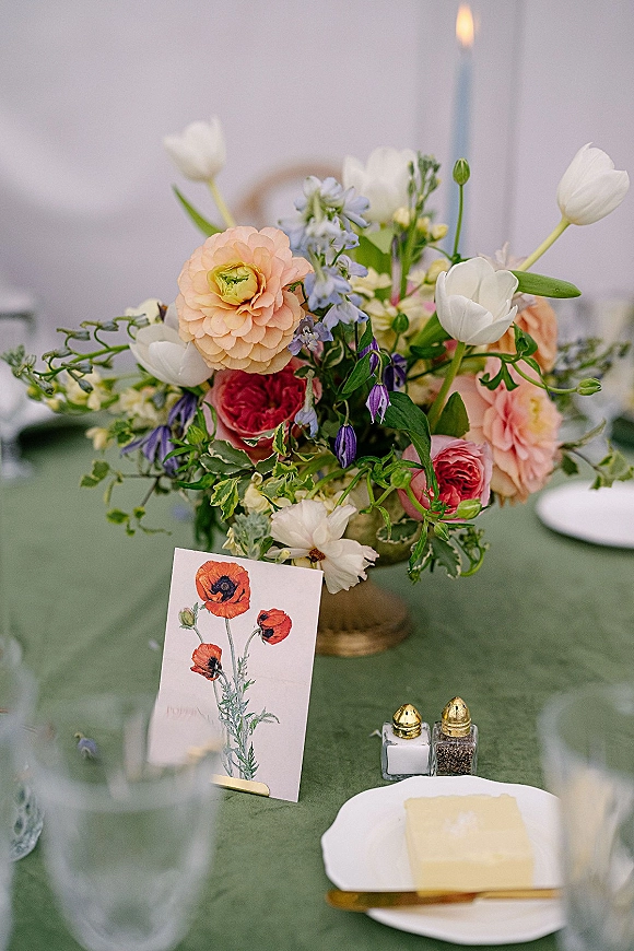Reception tablescape with a wedding table centerpiece in a pedestal vase, taper candle, place card, gold flatware, and sage linen
