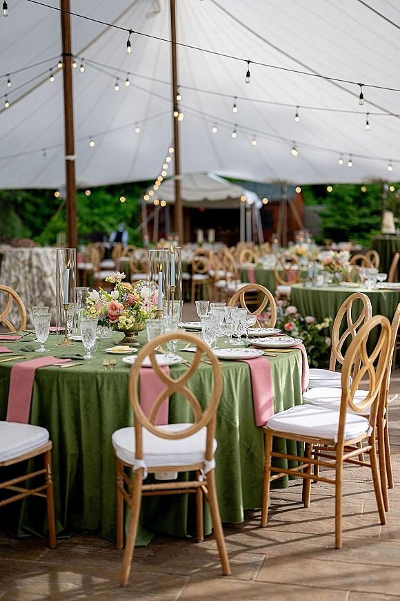 Reception tablescape with green table linens and a pink runner, floral centerpieces and candles, set under a white tent with string lights