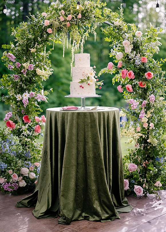 Wedding cake display with a three tier wedding cake on a stand, rose topper and floral arch, set on a green velvet table outdoors