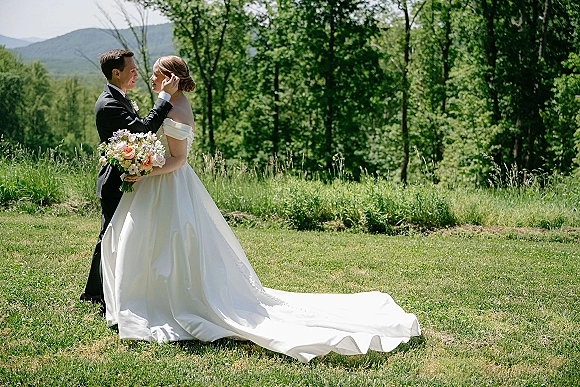 Couple portrait of bride and groom embracing, groom touching her face as she holds a bouquet in a mountain meadow with trees