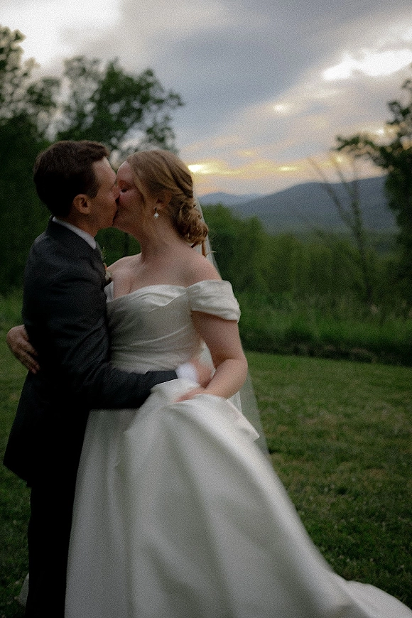 Wedding kiss as the bride and groom kiss in a grassy field, her long veil in sunset light with mountains and a cloudy sky behind