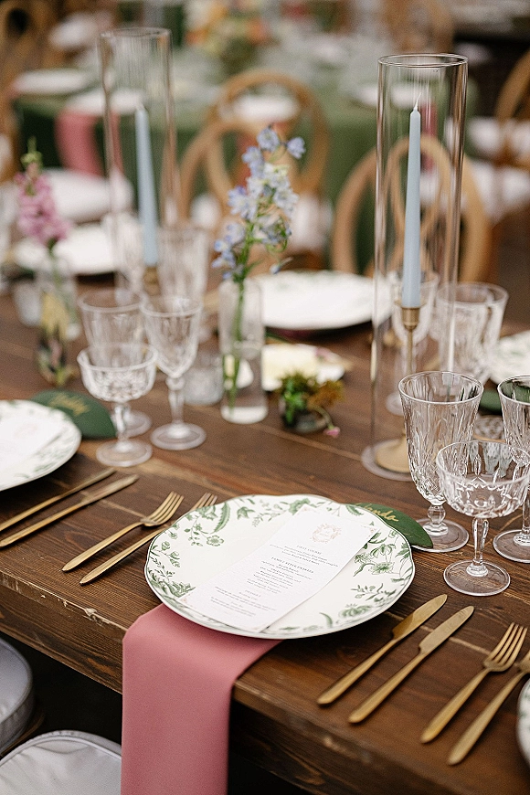 Reception tablescape with botanical wedding place setting, gold flatware, crystal goblets, blush runner, taper candles and bud vases on a wood table
