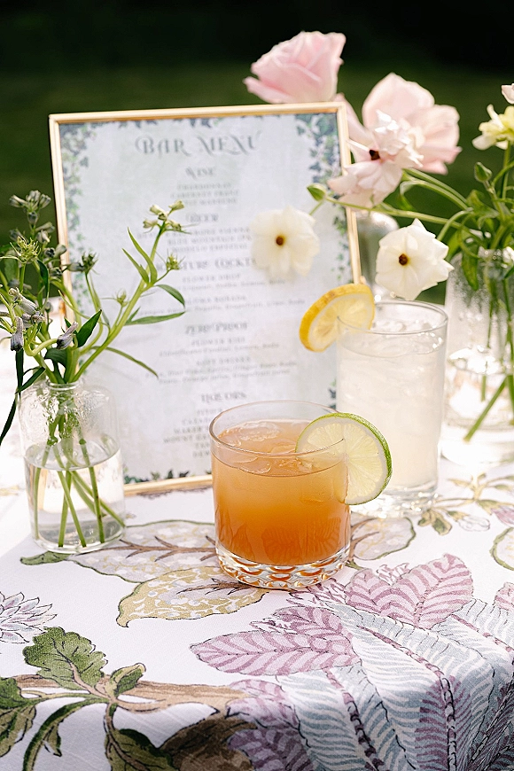 Wedding menu sign in a gold frame with floral border, bud vases, and cocktails with lime and lemon garnish on a patterned table outdoors