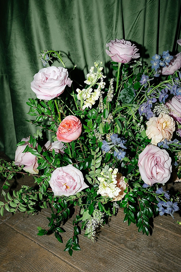 Wedding floral arrangement with garden roses, blue delphinium, and greenery on a wooden floor against a green draped backdrop