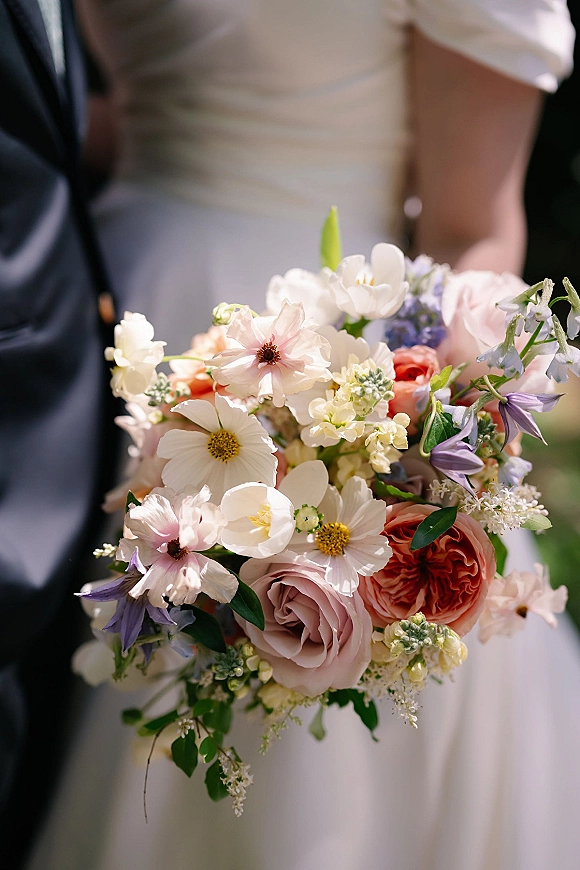 Bridal bouquet, a pastel wedding bouquet of garden roses and cosmos with greenery, held in sunlight with a blurred couple in the garden background