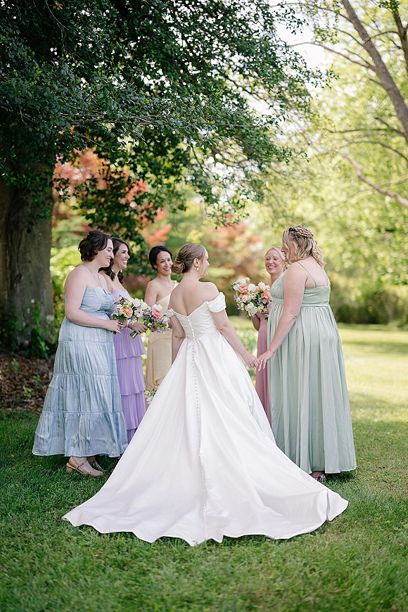 Bridesmaids portrait with bride in off-the-shoulder gown, pastel dresses and bouquets on a green lawn under a large leafy tree