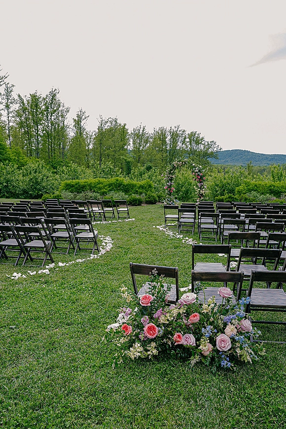 Outdoor ceremony setup with a floral arch, wood folding chairs in a circular layout, and white aisle petals on a lawn with hills beyond