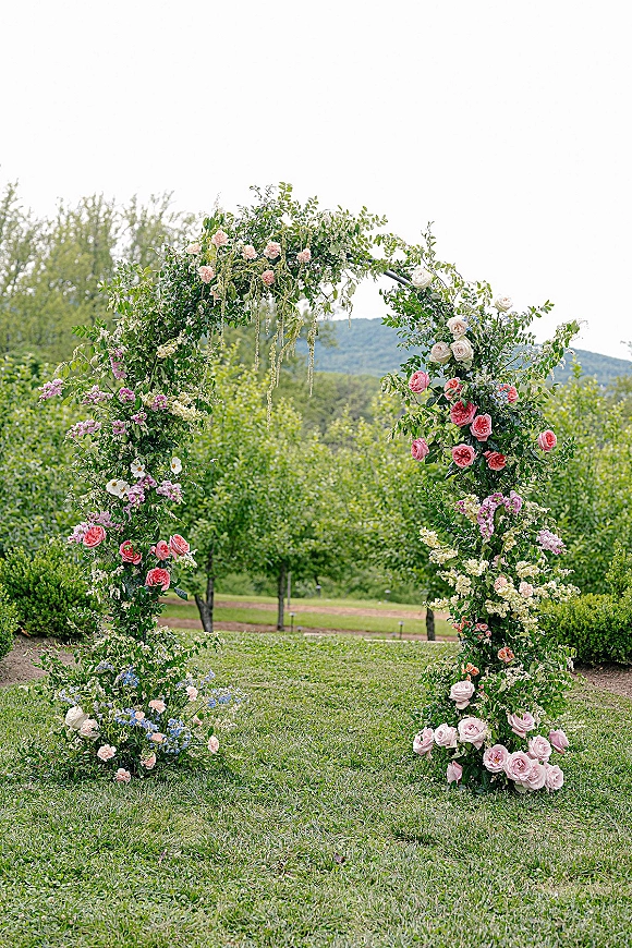 Wedding ceremony arch with a circular frame draped in greenery and pastel rose clusters on a lawn with orchard trees and hills behind