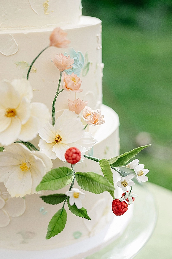 Wedding cake with floral wedding cake sugar flowers, buttercream frosting, and red berry accents on a stand against blurred greenery