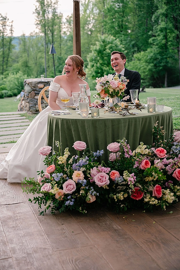 Reception moment as bride and groom laughing at a sweetheart table with candles and floral centerpiece on an outdoor patio lawn