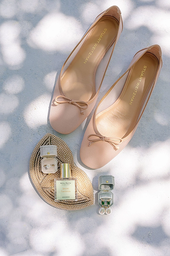Bridal accessories flatlay with blush ballet flats, pearl heart clutch, diamond ring, bands, perfume bottle, and earrings on white surface