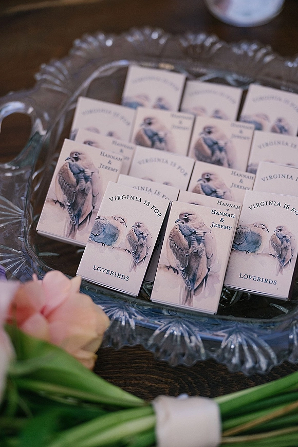 Wedding favors featuring custom matchboxes wedding with bird illustration labels on a glass platter on a wood table with a pink rose