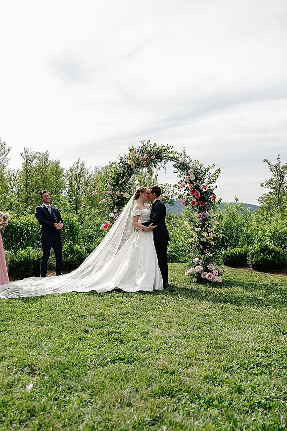 Wedding kiss at an outdoor ceremony kiss beneath a rose-and-greenery floral arch, bride in off-the-shoulder gown and long veil on garden lawn