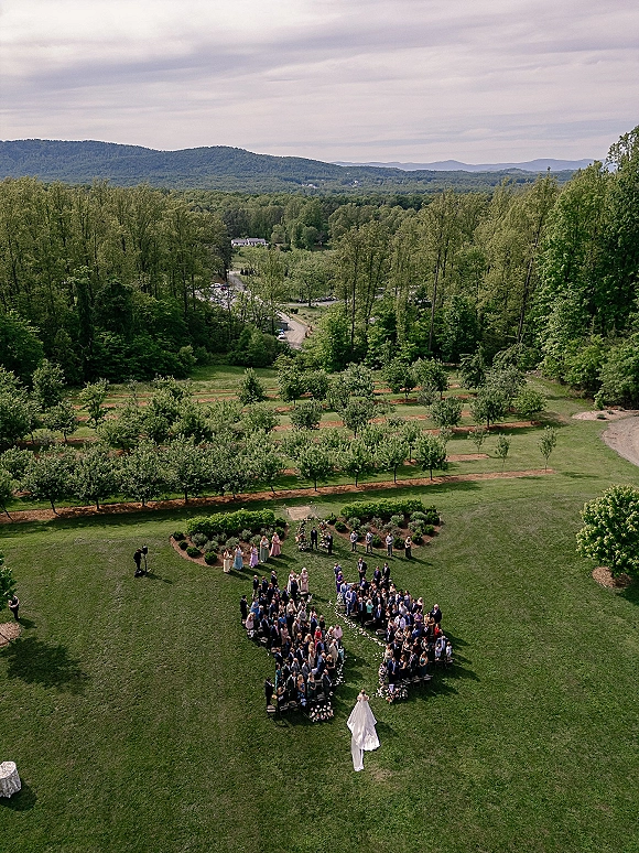 Outdoor ceremony moment in an aerial wedding ceremony with a flower petal aisle, rows of chairs, and mountains under a cloudy sky