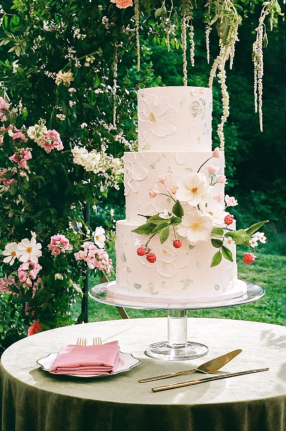 Wedding cake with buttercream floral piping and sugar flowers, raspberry accents, displayed on a stand on a garden table under blooms