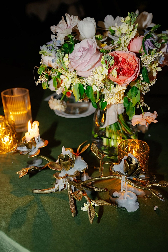 Reception tablescape with a wedding table centerpiece of pink and coral roses in a glass vase, taper candles and amber votives in a dark room