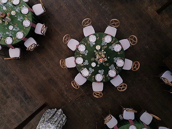 Reception tablescape overhead with round wedding tables in green tablecloths, gold flatware, patterned plates, floral centerpieces and candles on dark wood floor