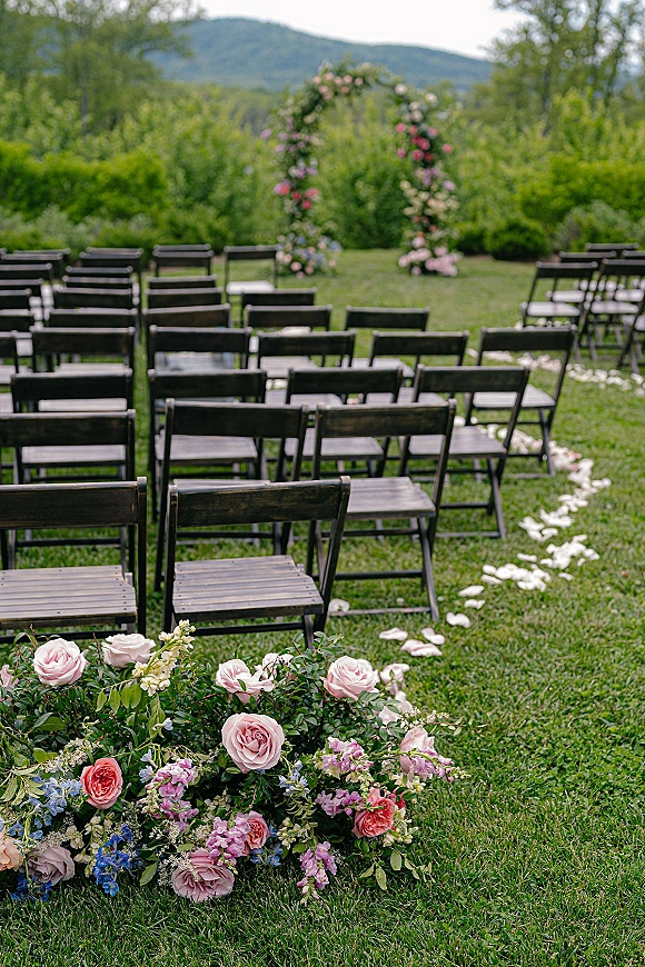 Outdoor ceremony setup with wood folding chairs and rose petal aisle leading to a floral arch on a lawn with mountain views