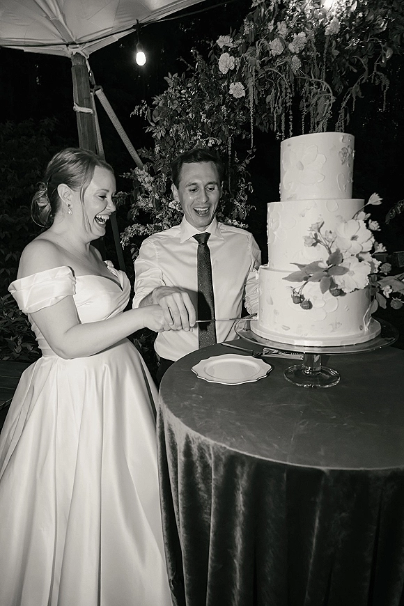 Cake cutting moment as bride and groom hold the knife over a three-tier buttercream cake on a stand beneath string lights in a tent at night