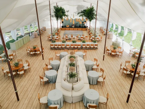 Wedding reception setup under a white tent reception with round tables, banquet table, greenery centerpieces, lantern lights, stage, and wood dance floor
