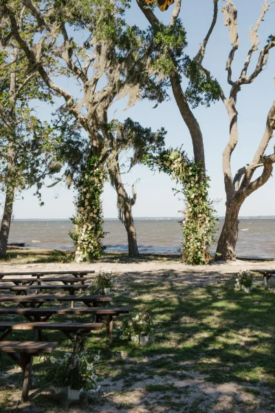 Ceremony setup with a floral arch and greenery, white aisle florals, and wooden benches beneath oak trees by the ocean backdrop