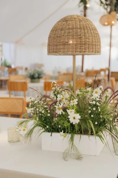 Reception tablescape with a wedding table centerpiece of wildflowers, white daisies and greenery, plus a rattan lamp under tent string lights