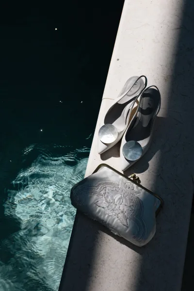 Bridal accessories with white slingback heels and embroidered satin clutch, styled by sunlit pool water and stone edge shadows