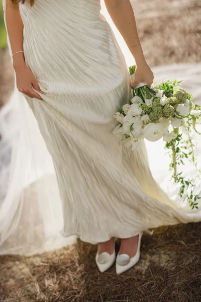 Bridal gown details in a ruched wedding dress as the bride holds a white-and-green bouquet in sunlight with veil, bracelet, and pumps