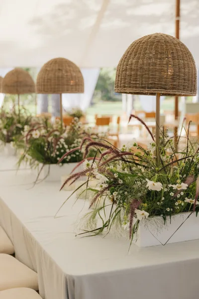 Reception centerpiece decor with a rattan table lamp centerpiece, white flowers and pampas in planter boxes on a long table under a white tent canopy