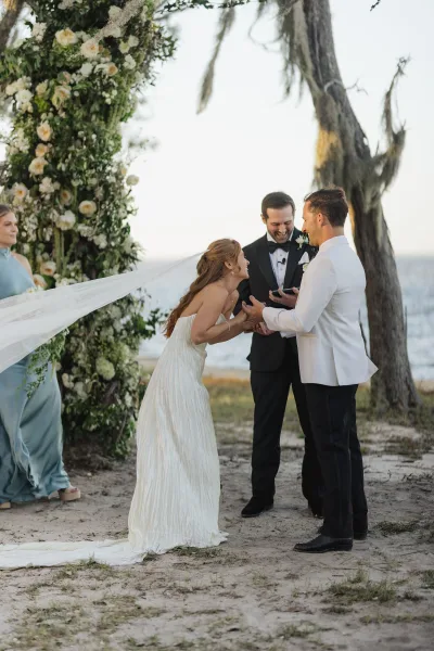 Ceremony moment as bride and groom exchange vows holding hands under a floral arch, with ocean shoreline and a large tree behind them