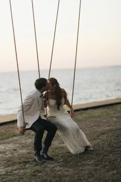 Wedding kiss portrait of bride and groom kissing on a rope swing, her strapless gown and his tuxedo against ocean shoreline at sunset