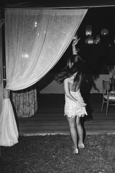 Wedding dance moment of a bride dancing in a short lace open-back dress, one drink held high, under lantern-lit tent at night