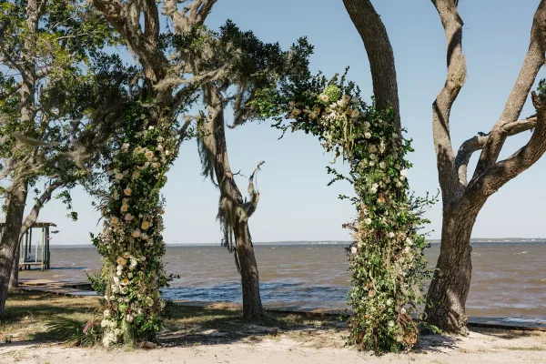 Wedding ceremony arch with floral arch greenery and white roses, framed by oak branches and Spanish moss on a waterfront shoreline under blue sky