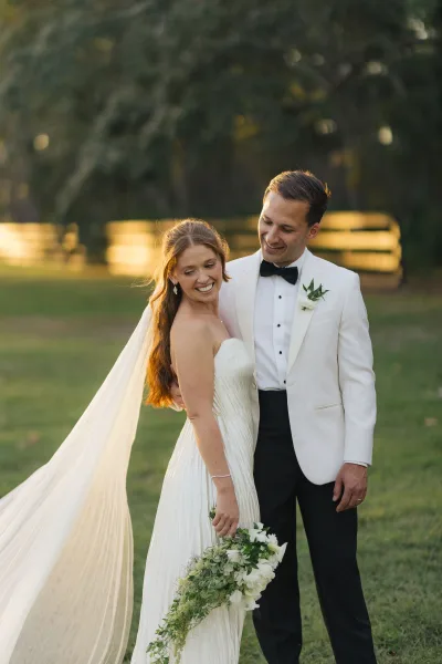 Couple portrait of bride and groom embracing on a grassy lawn, her veil blowing as she holds a cascading bouquet by a wooden fence