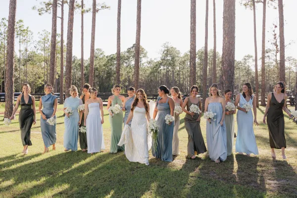 Bride with bridesmaids walking together, holding white bouquets with greenery accents in mismatched dresses across a grassy field by pine trees