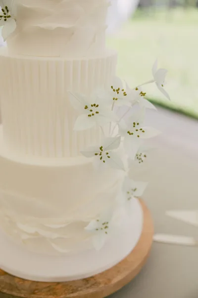 Wedding cake with fluted buttercream and white sugar flowers on a stand, set on a table with soft outdoor greenery blurred behind