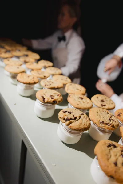 Wedding dessert table with chocolate chip cookies and milk jars arranged for grab-and-go treats, with caterers against a dark backdrop