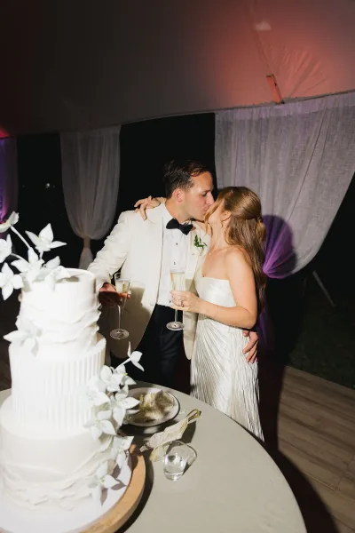 Wedding cake moment as bride and groom share a cake kiss beside a white floral cake, raising champagne flutes under a draped tent at night