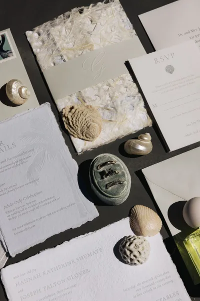 Invitation suite with textured handmade paper and deckled edges, styled with wax seal, rings, seashells, and perfume on a dark tabletop