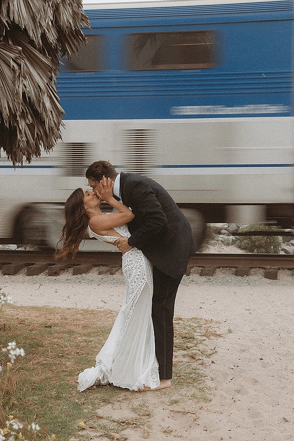 Wedding kiss portrait of groom dipping the bride in a lace dress beside railroad tracks as a train blurs past in the background
